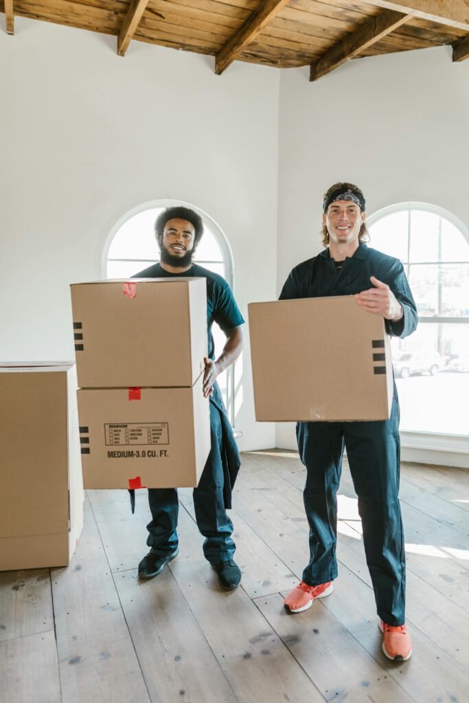 Two smiling movers carrying cardboard boxes in a bright, airy room with wooden floors.